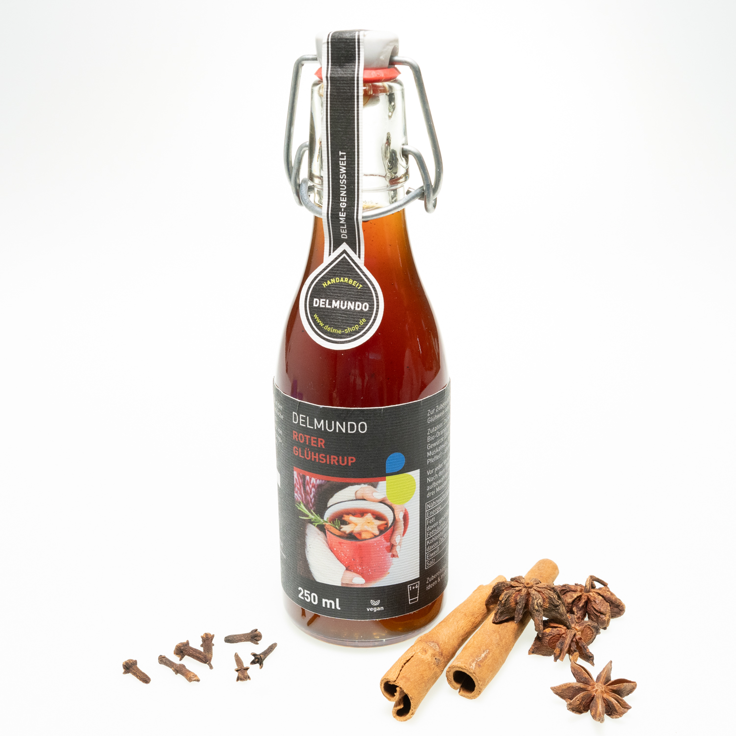 Eine klare Glasflasche mit Bügel-Verschluss gefüllt mit kräftig rot-braunem Glühsirup. Daneben liegen Zimtstangen und Gewürznelken.