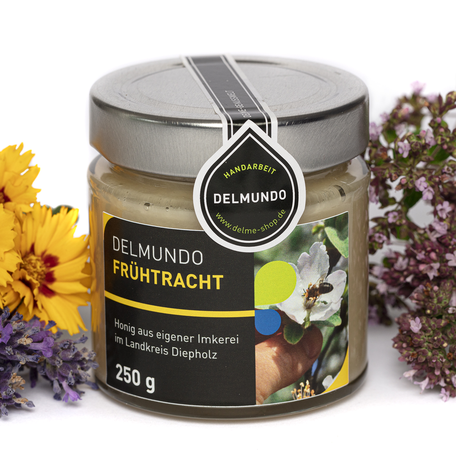 Rundes Glas mit silbernem Deckel, gefüllt mit hellem cremigen Frühtracht-Honig. Daneben liegen diverse blühende Blumen.