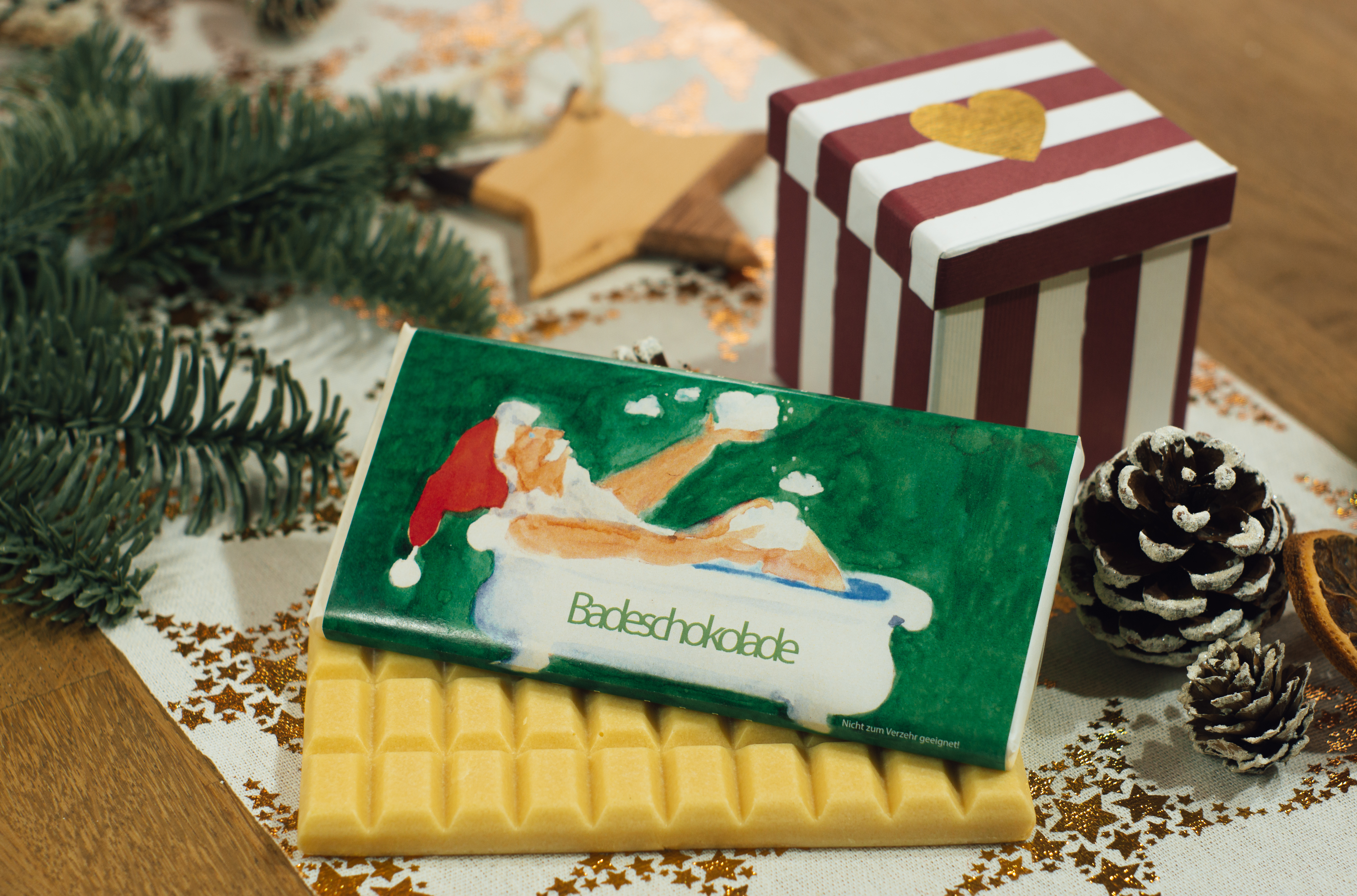 Cremeweißer Badezusatz in Schokoladenform. Auf der Packung der Badeschokolade ist ein gemalter Weihnachtsmann in einer Badewanne vor einem dunkelgrünem Hintergrund abgebildet.