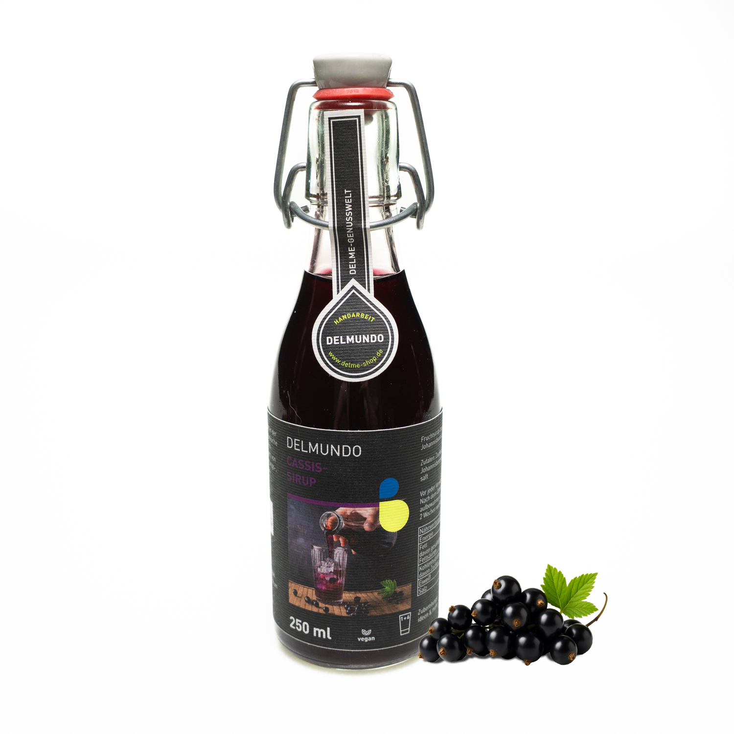 Eine klare Glasflasche mit Bügel-Verschluss gefüllt mit dunklem Cassis-Sirup. Daneben liegt ein Strauch Cassisbeeren.