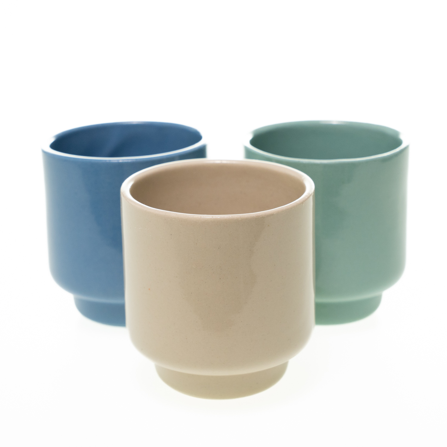 Drei schlichte, zylindrische Keramikbecher in mit glatter Oberfläche in den Farben blau, mintgrün und beige. Der untere Rand ist leicht zurückgesetzt.