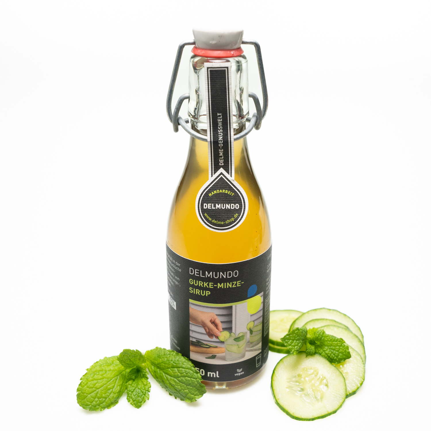 Eine klare Glasflasche mit Bügel-Verschluss gefüllt mit erfrischend grün-goldenem Gurke-Minz-Sirup. Daneben liegen Gurkenscheiben und Minzblätter