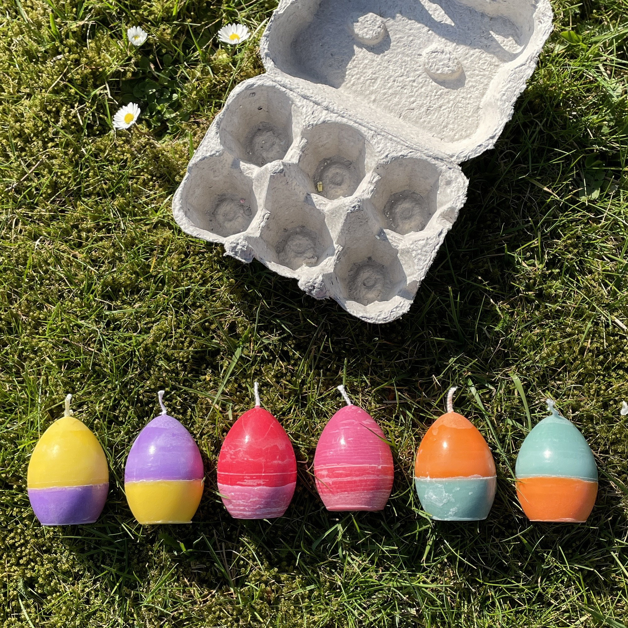 6 verschiedenfarbige Kerzen in Form eines Eis in einem Eierkarton in den Farben: Gelb-Lila,  Lila-Gelb, Rosa-Rot, Rot-Rosa und Orange-Grün, Grün-Orange. Die Teilung des verschiedenfarbigen Wachses ist gerade durch die Mitte.
