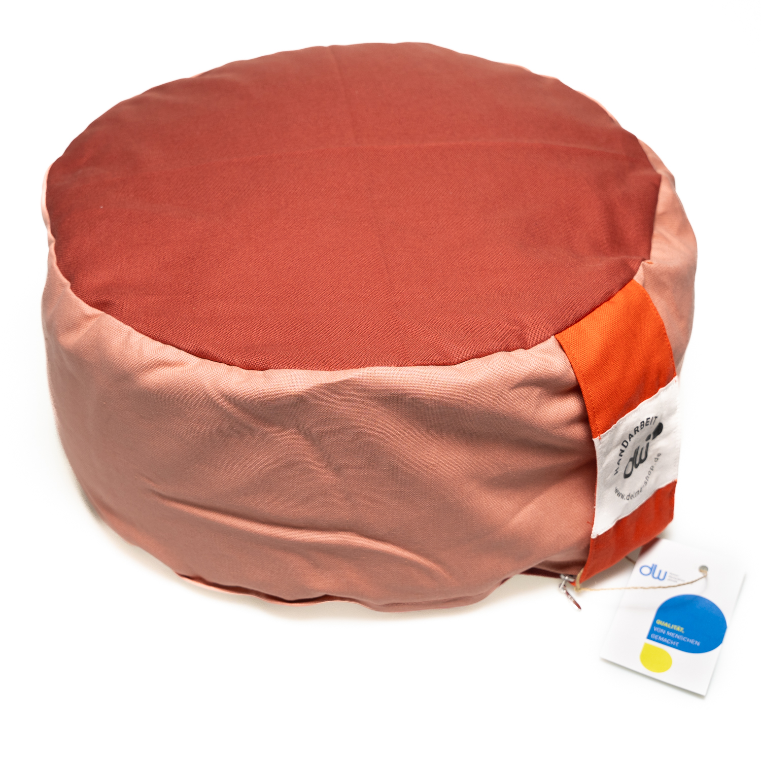 Rundes Yogasitzkissen. Die Seite ist rosé oben und unten ist der Stoff terakottafarben. An der Seite ist eine orangene Stofflasche zum Tragen und der Bezug kann mit einem Reißverschluss geöffnet werden.