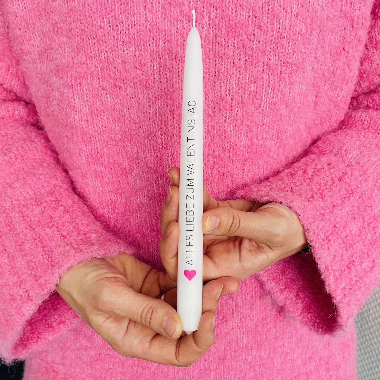 Weiße Tafelkerze mit der Aufschrift „Alles Liebe zum Valentinstag“ in schlichten schwarzen Versalien geschrieben. Unten am Anfang des Satzes befindet sich ein kleines pinkes Herz.  Die Kerze wird von zwei Händen gehalten