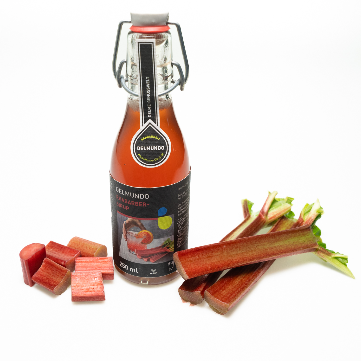 Eine klare Glasflasche mit Bügel-Verschluss gefüllt mit tiefrotem Rhabarbersirup. Daneben liegen Rhabarberstücke und ganze Rhabarberstangen.